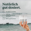 Hand hält ein Glas Wasser mit Informationen zur Dosierung von Nature Love Premium Collagenpulver vor einem Meereslandschaftshintergrund.